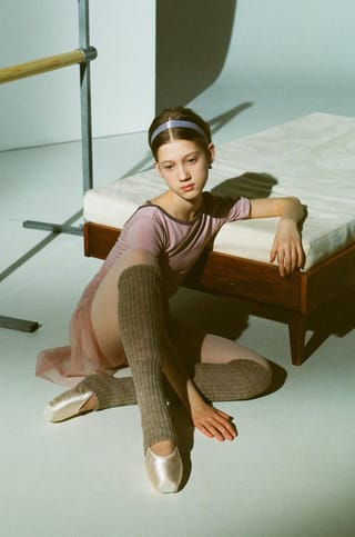 Bir Çocuk pink leotard ve tül tülleri giymiş olarak bir bale stıdyosunda, arka planda bir yatak ve bale barre var.