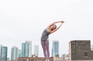 A woman in yoga attire performing a stretch on a rooftop with a cityscape behind her.