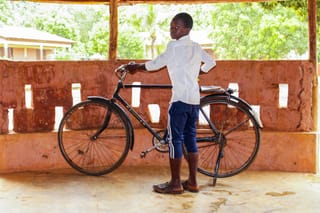 A young boy in a white shirt and blue shorts stands with a bicycle in a rustic outdoor setting.