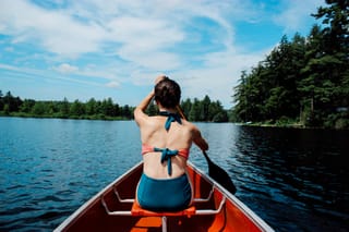 Back view of a woman in a bikini paddling a red canoe on a calm lake with a forest in the background.