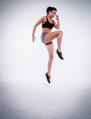 A woman in a black sports bra and grey shorts leaps in the air, wearing black sneakers with pink laces.