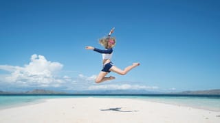 A woman in a blue and white swimsuit mid-jump on a white sandy beach with a clear blue sky and ocean in the background.
