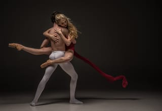 A male dancer in white tights lifts a female dancer in a red dress against a dark background.
