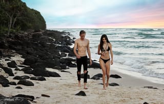 A shirtless man in black pants and a woman in a black bikini walking side-by-side on a beach with black rocks and the ocean in the background.