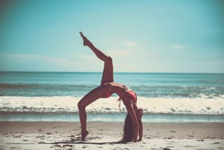 A woman in a pink bikini performing a standing backbend yoga pose on a sandy beach with ocean waves in the background.