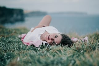 A woman with dark hair wearing a white top and pink skirt lying in green grass on a hillside, smiling, with a blurred ocean view behind her.