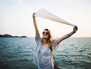 A woman with blonde hair and sunglasses smiles while holding a white scarf above her head with both hands, standing against a backdrop of the blue sea and distant islands.
