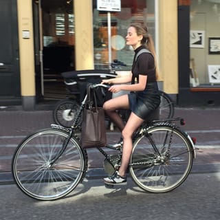 A woman cycling on a city street, wearing a black dress and carrying a brown bag.