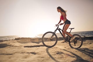A woman in a red top and black shorts riding a mountain bike on a beach with the ocean in the background.