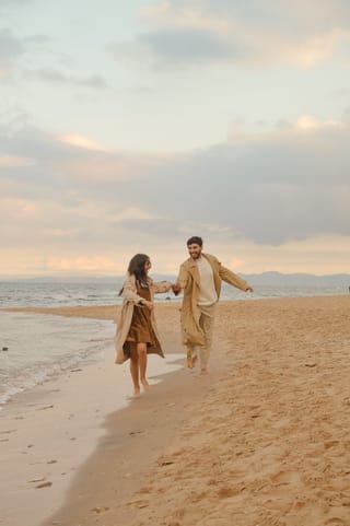 Two individuals, a man and a woman, running hand in hand on a sandy beach with the ocean in the background.