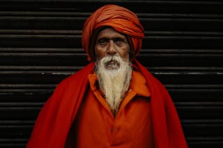 An elderly man with a red turban and beard, standing in front of a dark background.