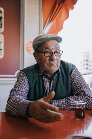 An elderly man with glasses and a flat cap sits at a red table in a cafe, gesturing with his hand. He is wearing a plaid shirt and a green vest. A glass of tea is on the table in front of him.