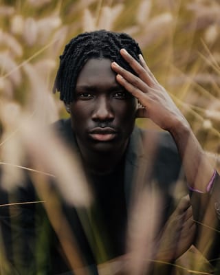A portrait of a young Black man with dreadlocks looking directly at the camera, with his hand resting on his forehead, set against a background of blurred tall grass.