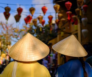 Back view of two people wearing traditional conical hats walking under strings of colorful lanterns.