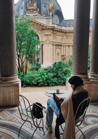 A person sitting at a cafe table under large stone columns, looking at a phone, with the ornate facade of the Musée d'Orsay in the background.
