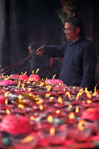 A man wearing glasses and a dark blue shirt stands in a temple, holding incense sticks over a large collection of lit red lotus-shaped candles.