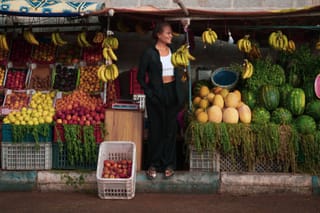 A woman standing in a fruit market stall surrounded by hanging bananas, melons, watermelons, and crates of apples and other fruits.