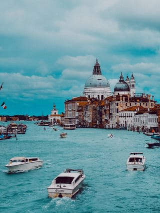A wide shot of the Grand Canal in Venice with the Basilica di Santa Maria della Salute in the background. Several white water taxis are moving through the blue-green water. The sky is overcast with blue and grey clouds.