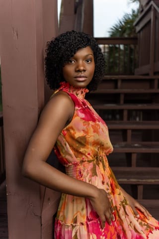 A portrait of a young woman with dark skin and short curly black hair, wearing a colorful floral halter-neck dress, leaning against a brown wooden post with stairs in the background.
