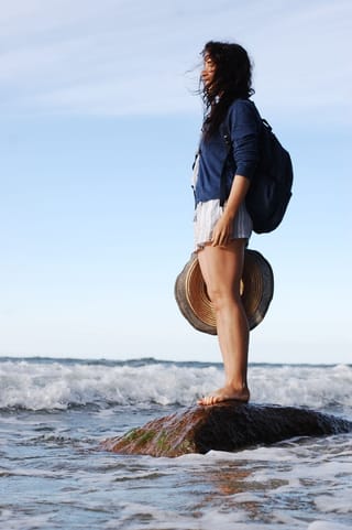 A woman standing on a rock in the ocean, holding a hat and wearing a backpack.