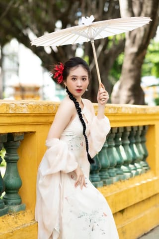Una mujer viste un traje tradicional blanco vietnamita con patrones florales, sosteniendo una sombrilla de papel blanco, parada junto a una barandilla de un puente amarillo y verde con árboles en el fondo.