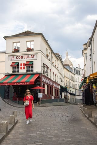 A woman in a red dress walking on a cobblestone street in front of the Le Consulat cafe in Montmartre, Paris, with the Sacré-Cœur Basilica visible in the background.