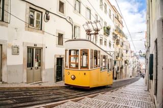 A yellow tram moving down a steep, narrow cobblestone street flanked by white buildings with balconies.