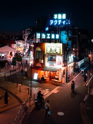 A night scene of a Japanese street corner with glowing neon signs, including one for a real estate agency, and people walking on the sidewalk.