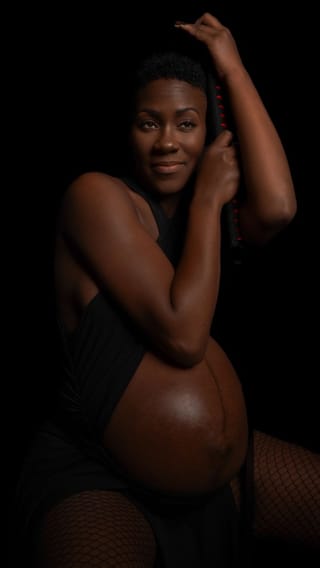 A pregnant woman with short hair poses against a black background, wearing a black top and fishnet stockings, holding a red beaded accessory.