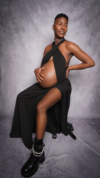 A pregnant woman with short hair sits on a stool, wearing a black halter dress that exposes her belly, fishnet stockings, and black combat boots with pearl chains.