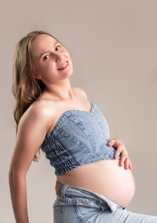 A pregnant woman with long blonde hair, wearing a strapless denim top and unbuttoned jeans, poses with her hand on her belly, looking back with a smile.