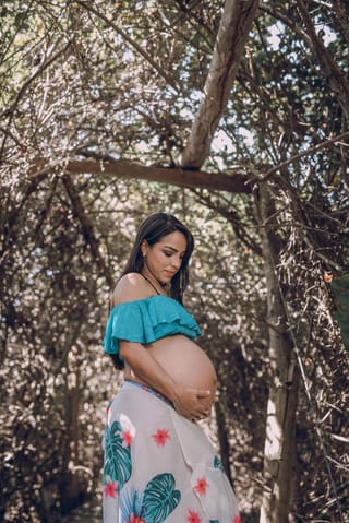 A pregnant woman standing outdoors in a forest setting, holding her baby bump. She is wearing a teal ruffled top and a white skirt with green leaves and pink flowers.