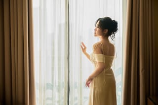 A woman in a yellow dress stands by a window with sheer curtains, looking outside.