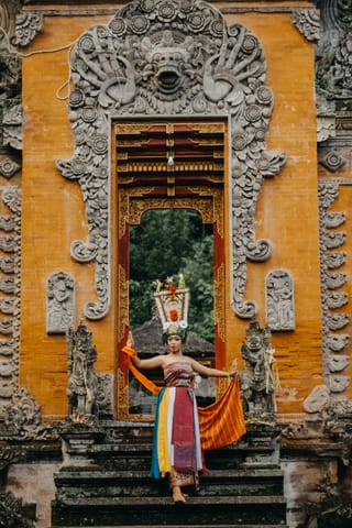 A woman in traditional Balinese dance costume stands on the steps of a highly decorated yellow temple gate, holding a colorful sash.