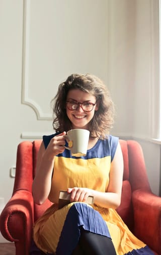 A woman with curly hair and glasses, wearing a yellow and blue dress, sits in a red armchair holding a white mug and a small book, smiling at the camera.