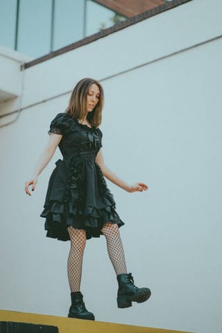 A young woman with shoulder-length brown hair stands against a white wall. She is wearing a black short-sleeved dress with ruffles, black fishnet stockings, and black platform boots. She is looking down and to her right.