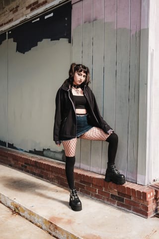 A young woman with dark hair in pigtails stands against a grey wooden wall and brick ledge. She is wearing a black oversized jacket over a black crop top, a denim mini skirt, fishnet tights, and black platform boots.