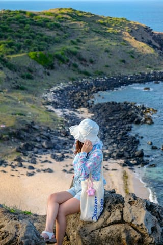 A woman wearing a white sun hat and a blue floral shirt sits on a large rock overlooking a coastal landscape with a sandy beach, dark volcanic rocks, and green hills meeting the blue ocean.