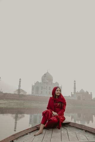 A woman in a red outfit sitting on a boat with the Taj Mahal in the background on a foggy day.