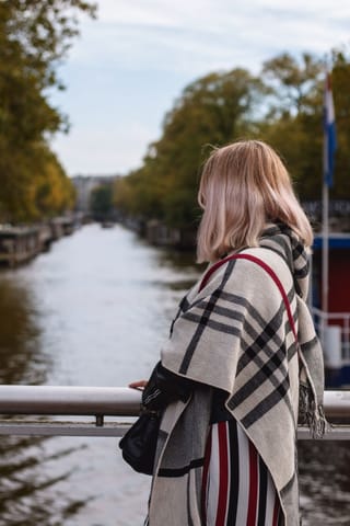 A woman with blonde hair wearing a plaid shawl stands by a canal, looking out over the water.
