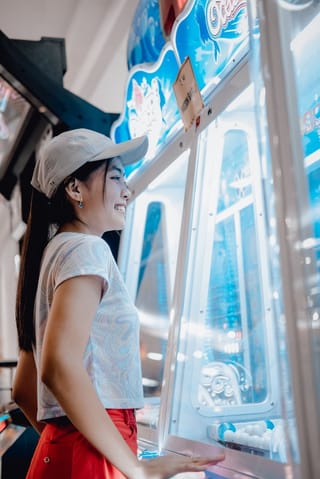 A young woman in a white cap and red shorts smiles while playing a claw machine arcade game.