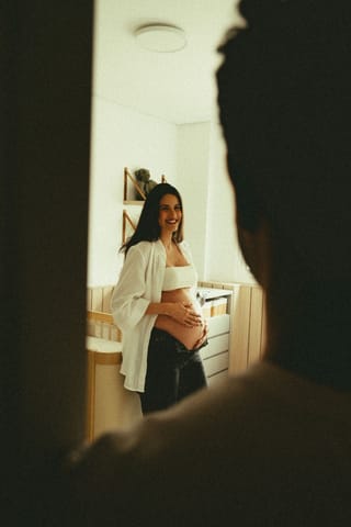 A pregnant woman wearing a white top and open shirt stands in a nursery, smiling and cradling her baby bump, seen through a doorway with a person's silhouette in the foreground.