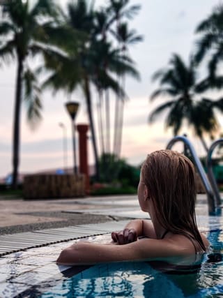 A woman in a pool, looking at the sunset, with palm trees in the background.