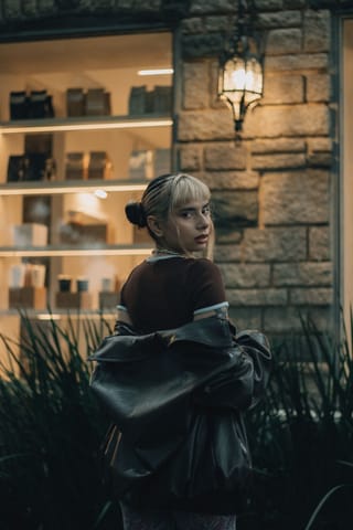 A young woman with blonde hair and bangs looking back over her shoulder, holding a black leather jacket, standing in front of a stone building with a lit window display and a wall lantern.