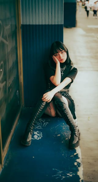 A young woman sitting on the ground, leaning against a wall, wearing a black dress and boots.
