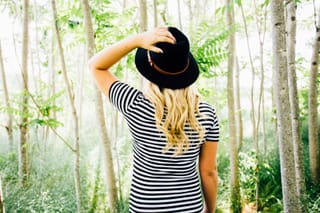 Woman wearing a striped shirt and black hat standing in a sunlit forest