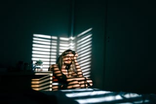 Woman seated indoors with dramatic striped shadows from window blinds