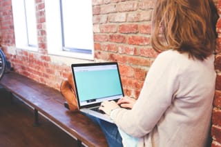 Woman working on laptop by brick wall