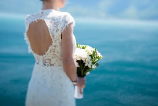 Bride in a lace wedding dress holds a bouquet while overlooking the blue sea