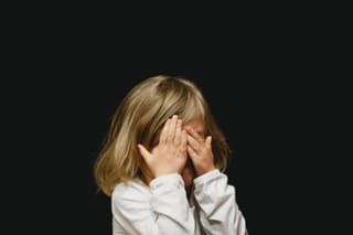 Young child covers their face with both hands against a dark background